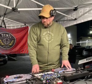 A man in a green Bent Water Brewing sweatshirt, yellow hat , and large headphones, DJs under a pop tent